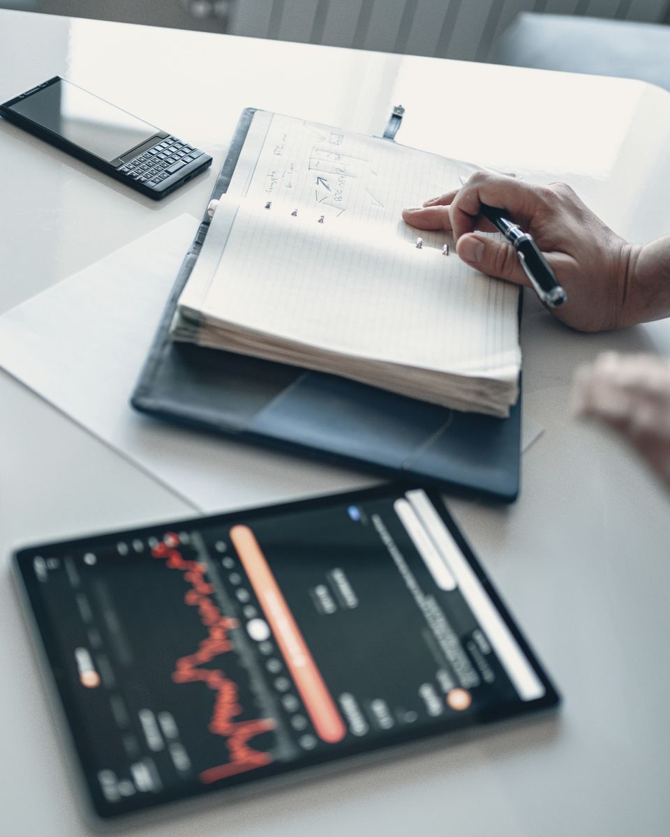 A hand writing in a notebook beside a tablet showing a performance chart.